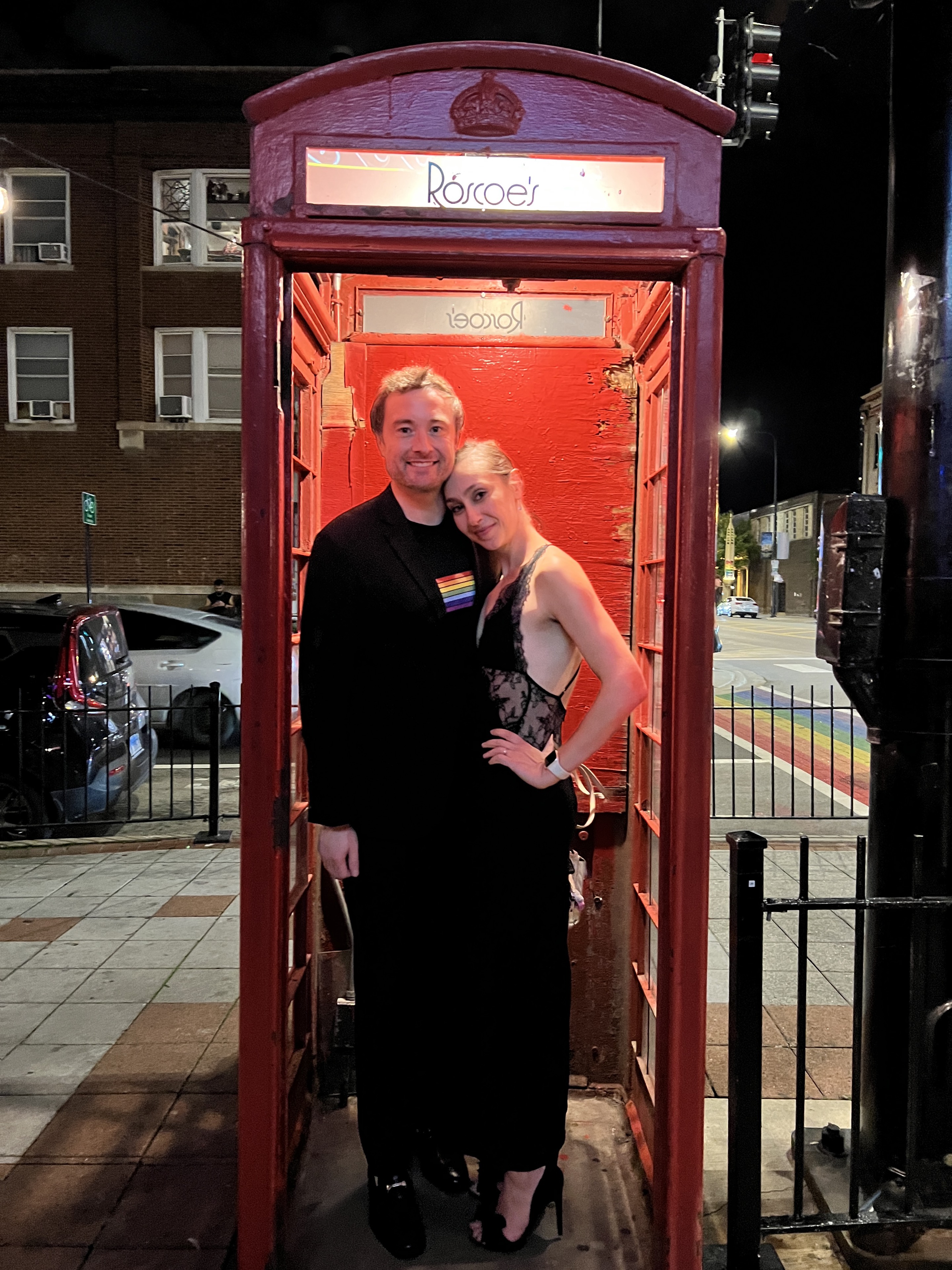 Two people posing together in a red telephone booth at night.